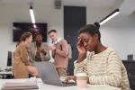 African-American woman working on a laptop alone at table with group of people whispering and bullying her in background. Visual concept for employment blog discussing the difference between workplace discrimination and harassment.